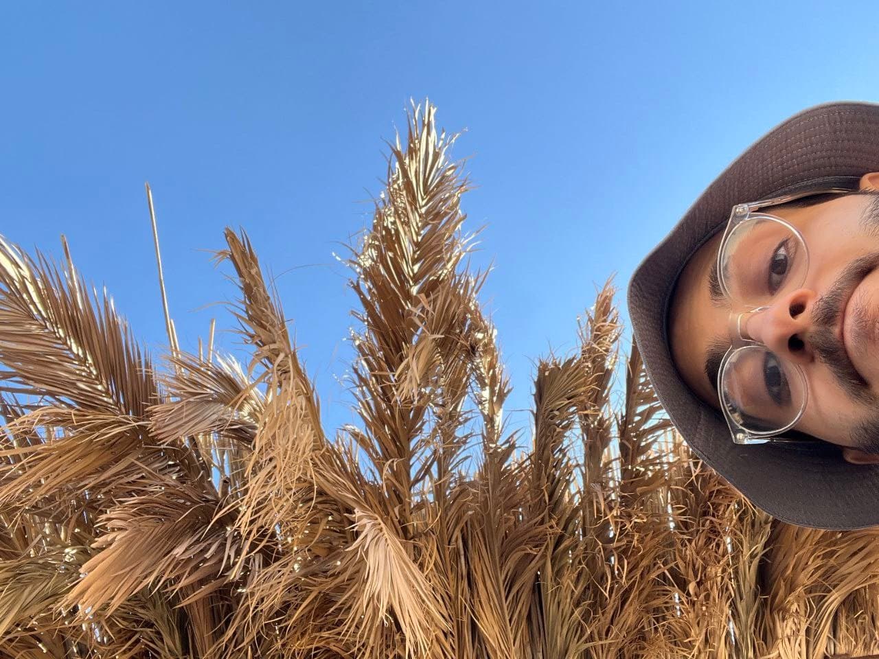 Kasra Hadizadeh in a dark bucket hat and clear round glasses, leaning into a selfie taken looking up, dried palm fronds against a clear blue sky beside him.